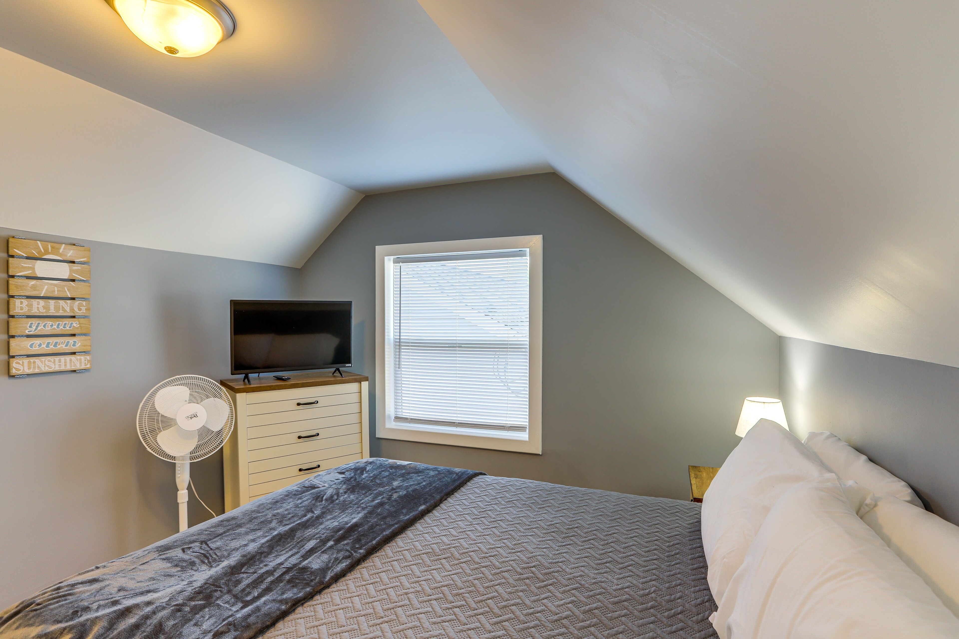 Ein Schlafzimmer mit Bett, Fernseher, Ventilator, Fenster mit Jalousien und Wandleuchte.