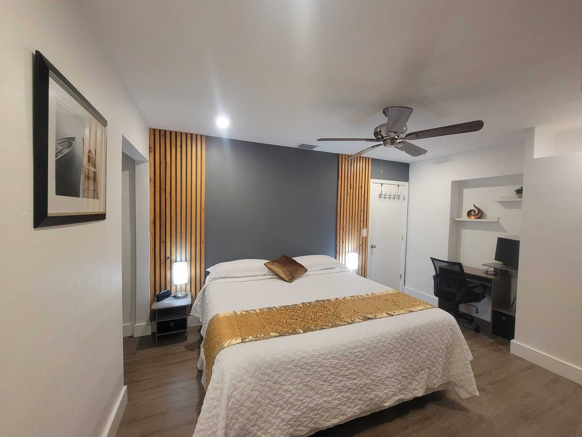 Un dormitorio con cama, escritorio, silla, ventilador de techo y puerta con perchero.