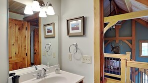 Un baño con un lavamanos blanco, tocador de madera y un espejo grande. Hay dos cuadros en la pared y un techo de madera con una decoración de cabeza de ciervo.