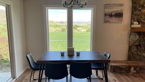 Uma sala de jantar com mesa e cadeiras, um lustre, uma parede de pedra e vista para uma paisagem verde através de uma grande janela.