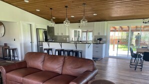 Uma sala de estar ampla com um sofá de couro marrom, uma mesa de centro de madeira e uma cozinha com armários brancos e uma ilha central.