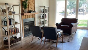 Uma sala de estar ampla com piso de madeira, lareira e duas poltronas de couro. Estantes repletas de livros e objetos decorativos.