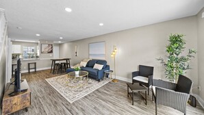 A modern living room with a blue sofa, a coffee table, and a floor lamp. There is a kitchen area with a window and a dining table. - Cozy Townhome| Moffitt| Pool| Gym| Fast WiFi| 2B2B (Tampa)