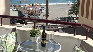 A balcony with a table, two chairs, a bottle of wine, and a glass. The view includes a beach, palm trees, and a building with a sign.
