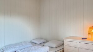 Een kleine eenpersoonskamer met witte wanden en een houten plafond. Er staat een nachtkastje met een lamp en een klein bordje op.