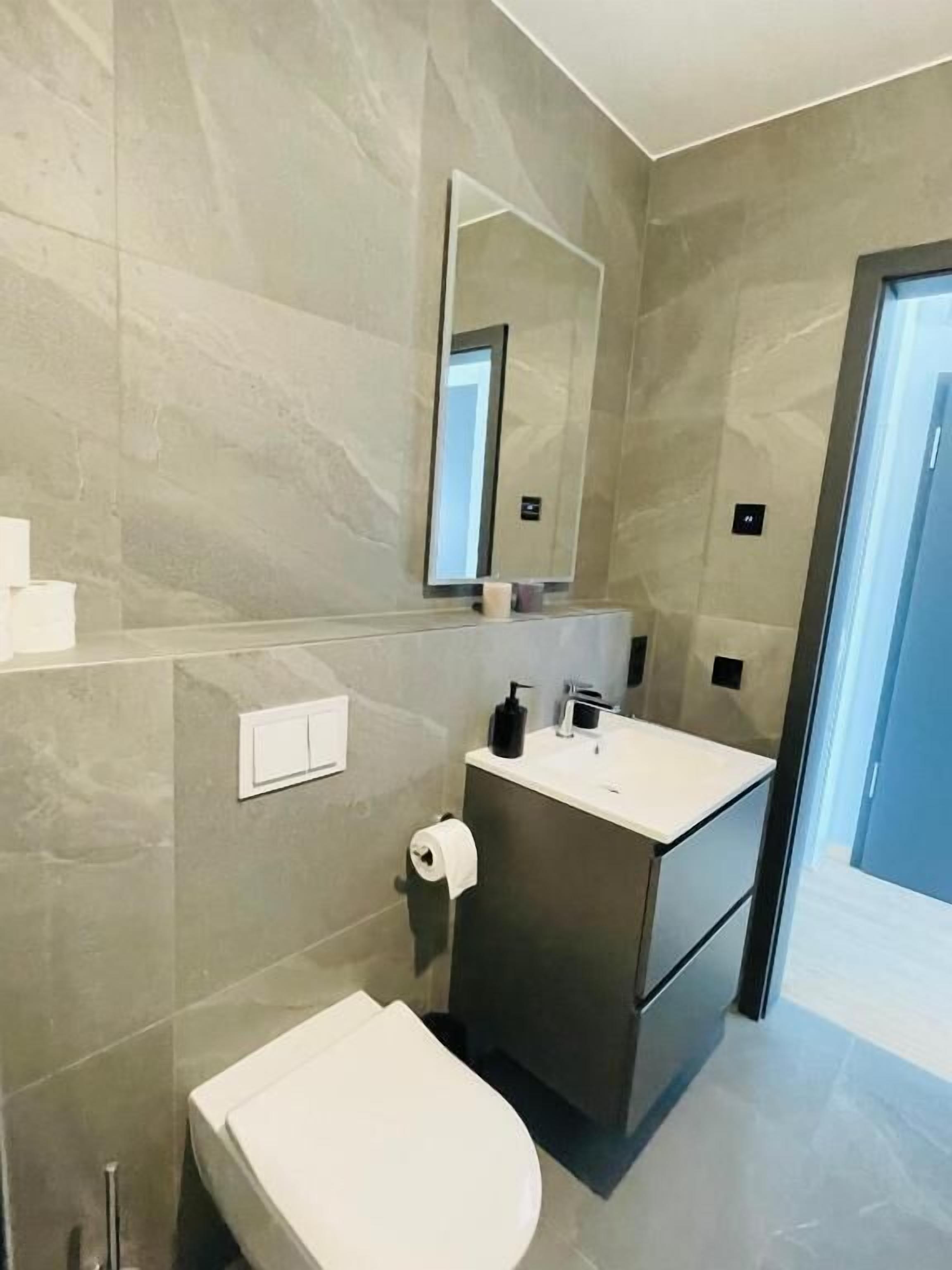A modern bathroom with a marble wall, a white toilet, a sink with a mirror above it, and a door leading to another room.