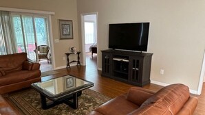 A living room with a brown leather sofa, a glass coffee table, and a flat-screen TV on a dark wooden stand. - 3 bedroom / 2 bath Courtyard Villa w/ cart! (The Villages)
