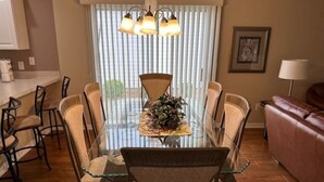 A dining area with a glass table surrounded by wicker chairs, a brown leather sofa, and a window with blinds. - 3 bedroom / 2 bath Courtyard Villa w/ cart! (The Villages)