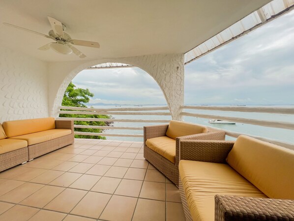 Een zonnig balkon met een plafondventilator, twee rieten banken en uitzicht op de zee.