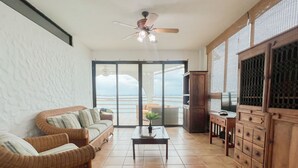 A living room with a sofa, a coffee table, a TV stand, and a view of the ocean through large glass doors. - Live an Adventure in Taboga 1 (Provincia de Panamá)