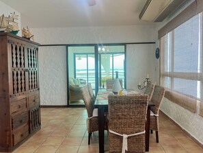 Een eetgedeelte met rieten stoelen, een houten kast en uitzicht op zee door een glazen deur.