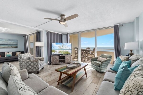 A spacious living room with a large TV, comfortable seating, a ceiling fan, and a balcony with a view.