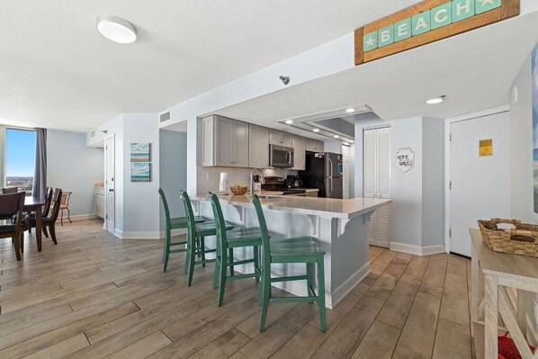 A modern kitchen with a central island, green chairs, and a view of the dining area. - Coastal Elegance Beachfront View (Pensacola)