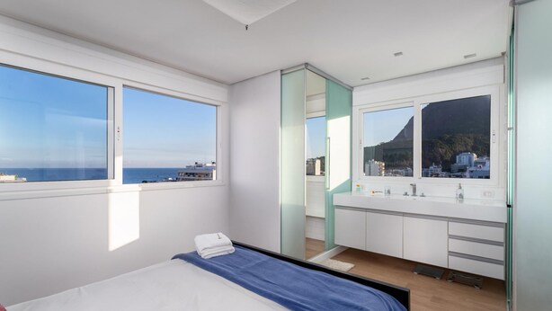A modern bedroom with a bed, a large window, a bathroom with a mirror, and a view of the sea. - Nice in Leblon View to Morro 2 irm os Guc6 ZN1 (Rio de Janeiro)