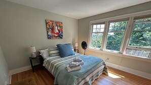 A bedroom with a bed, a bedside table, a floor lamp, a window with a view of trees, and a painting on the wall.