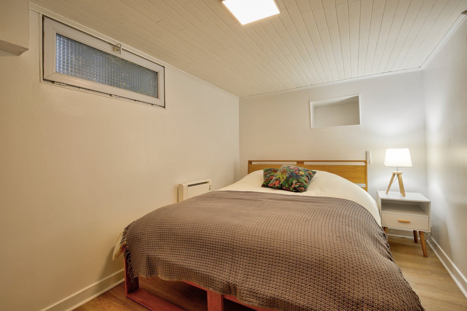 Une chambre à coucher avec un lit, une table de chevet, une lampe, une fenêtre et un luminaire au plafond.
