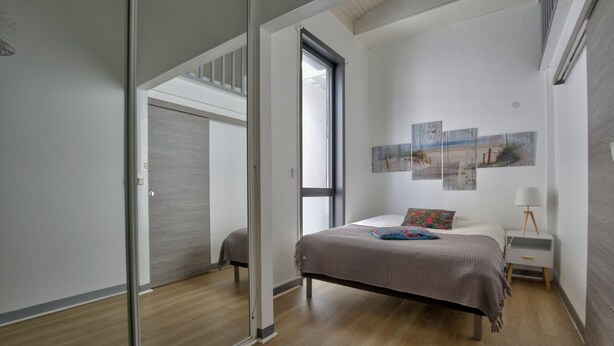 A modern bedroom with a bed, a nightstand, a lamp, and a large window. - Boulevard de la Plage flat-Arcachon (Arcachon)