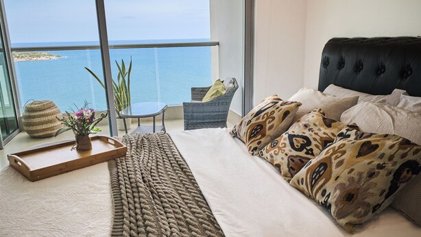 Ein Schlafzimmer mit großem Fenster mit Blick auf ein Gewässer, einem Bett mit gemusterten Kissen, einem Teppich und einem kleinen Tisch mit einer Pflanze.