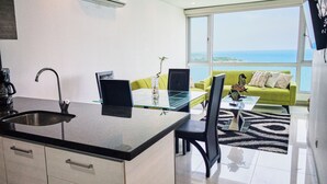 Eine moderne Küche mit einer Glasinsel, ein Essbereich mit einem grünen Sofa und Meerblick durch große Fenster.