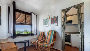Een kamer met twee gestreepte stoelen, een tafeltje, een raam met gordijnen, en een keuken die door een doorgang zichtbaar is.