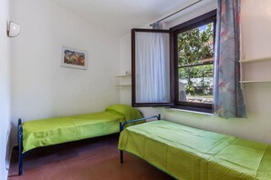 Une chambre avec deux lits simples, une fenêtre avec des rideaux, une petite étagère et un tableau encadré au mur.
