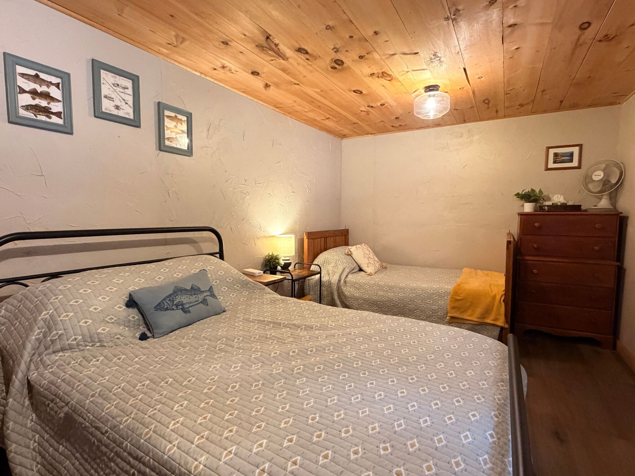 Un dormitori amb dos llits, sostre de fusta, quadres a la paret, una petita taula de nit, un aparador i un ventilador.
