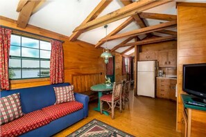 Ein gemütliches Blockhaus-Interieur mit Holzbalkendecke, einer blauen Couch mit gemusterten Kissen, einem Esstisch mit Stühlen, einer Küchenzeile mit Schränken und einem Kühlschrank sowie einem Fernseher auf einem Schrank.