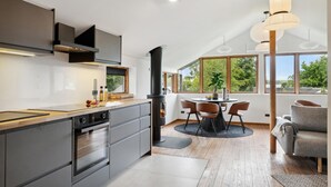 A modern kitchen with a central island, a dining area with a round table, and large windows offering a view of greenery. - 1 bedroom lovely home in Holbæk (Holbæk)