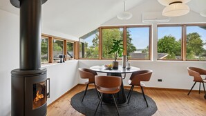 A modern living room with a wood-burning stove, a round dining table with chairs, and large windows offering a view of trees and buildings. - 1 bedroom lovely home in Holbæk (Holbæk)