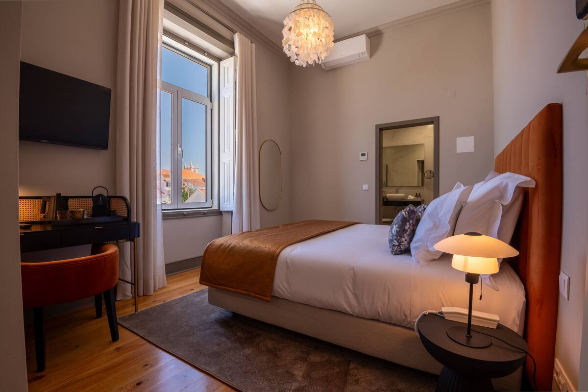 A modern hotel room with a bed, bedside table, lamp, desk, chair, and a TV.