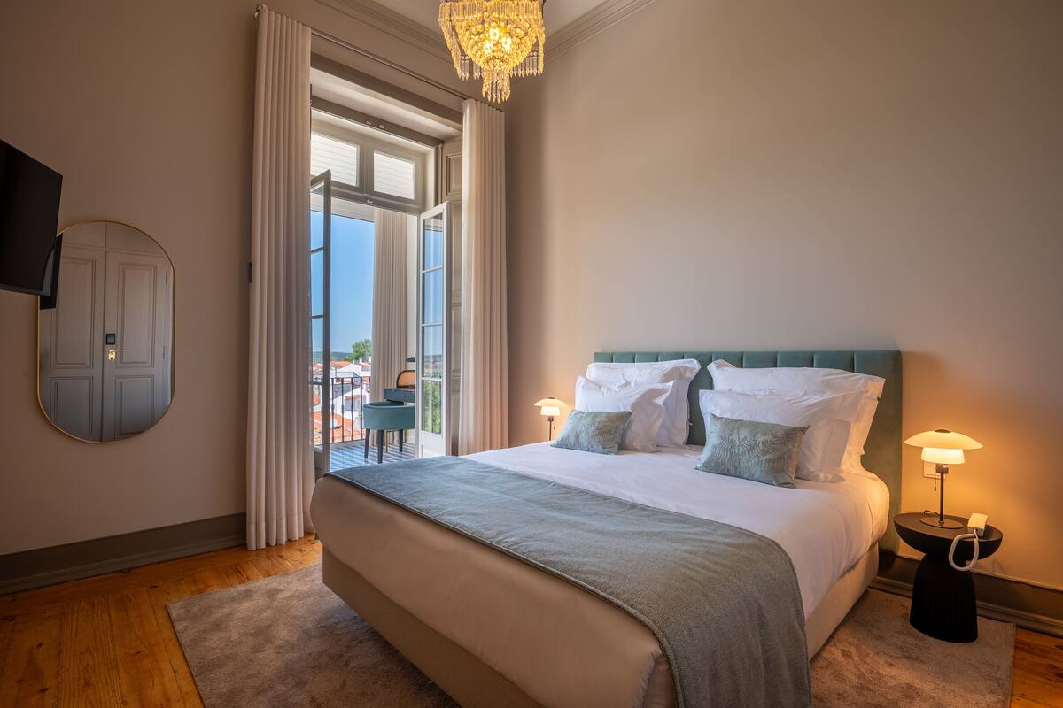 A bedroom with a large bed, a nightstand with a lamp, a TV mounted on the wall, and a view of the outdoors through a window.