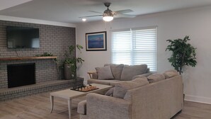 A living room with a brick fireplace, a flat-screen TV mounted above it, a beige sofa, a coffee table, and a ceiling fan. - 3 Bedroom House Near Hickory  (Granite Falls)