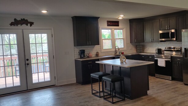 A modern kitchen with dark cabinetry, a central island with stools, a stainless steel refrigerator, and a window providing natural light. - 3 Bedroom House Near Hickory  (Granite Falls)