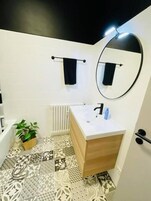 Un baño con piso de baldosas blancas, tocador de madera, lavamanos blanco, espejo y una planta en maceta.