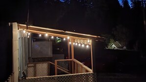 Un área de estar al aire libre con techo, luces colgantes y rodeada de árboles, con una casa a lo lejos.