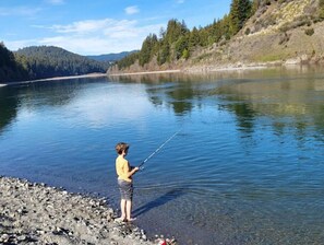 A person fishing by a calm lake with a rocky shoreline and forested hills in the background.