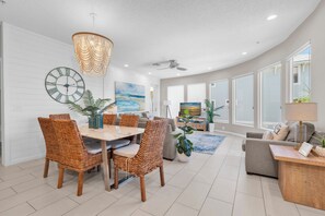 A modern living room with a dining area, featuring a large clock, wicker furniture, and a ceiling fan. - Beachfront Condo on AMI (Holmes Beach)
