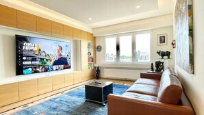 A modern living room with a large flat-screen TV mounted on the wall, a brown leather sofa, a blue patterned rug, and a window with a city view.