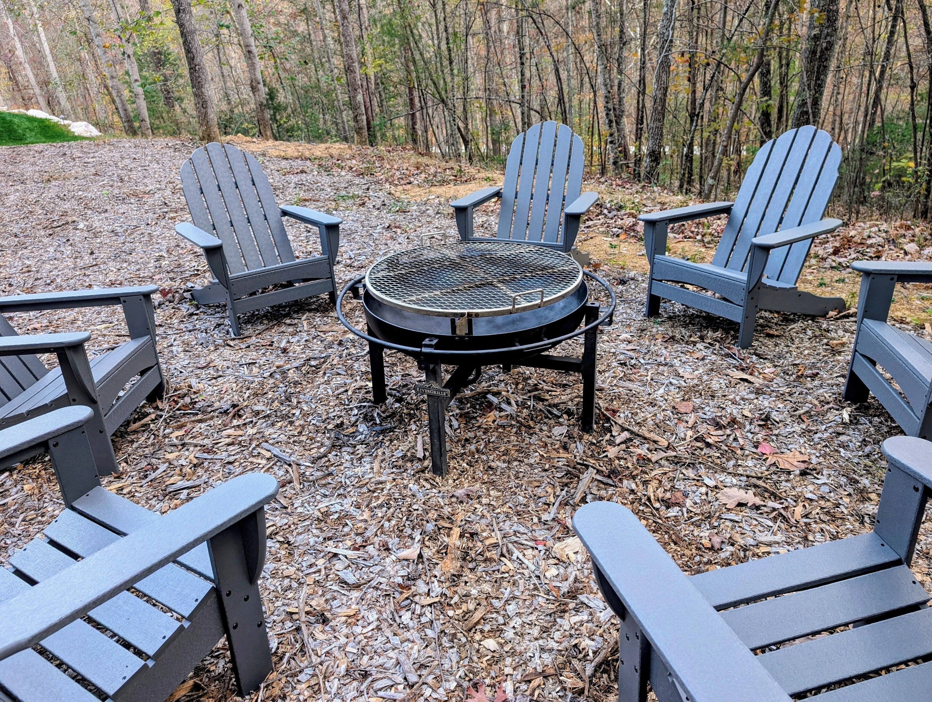 A circular outdoor grill surrounded by four Adirondack chairs in a wooded area.