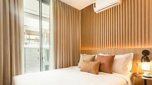 A hotel room with a large bed, a bedside table with a lamp, and a view through a window with blinds. - Helbor Wide (São Paulo)