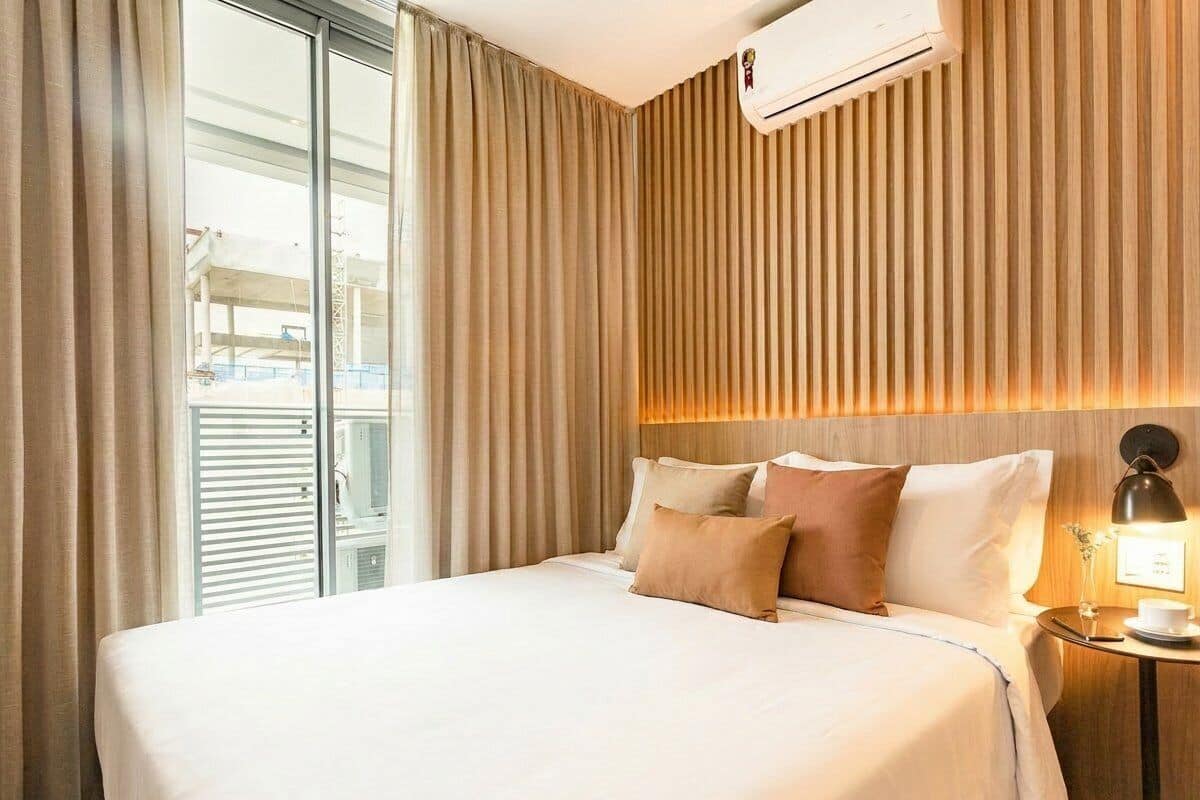 A hotel room with a large bed, a bedside table with a lamp, and a view through a window with blinds.