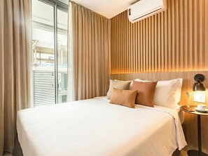 A hotel room with a large bed, a bedside table with a lamp, and a view through a window with blinds. - Helbor Wide (São Paulo)