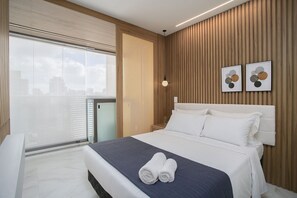 A modern hotel room with a large bed, a balcony with a city view, and a wooden accent wall. - Helbor Wide (São Paulo)
