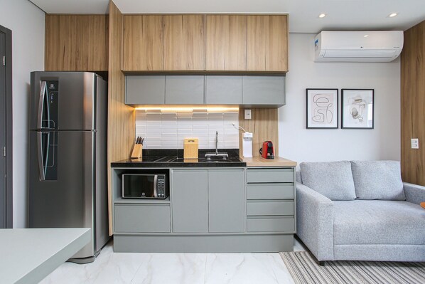 A modern kitchen with a built-in refrigerator, microwave, and stove, a grey sofa, and wooden cabinetry. - Helbor Wide (São Paulo)