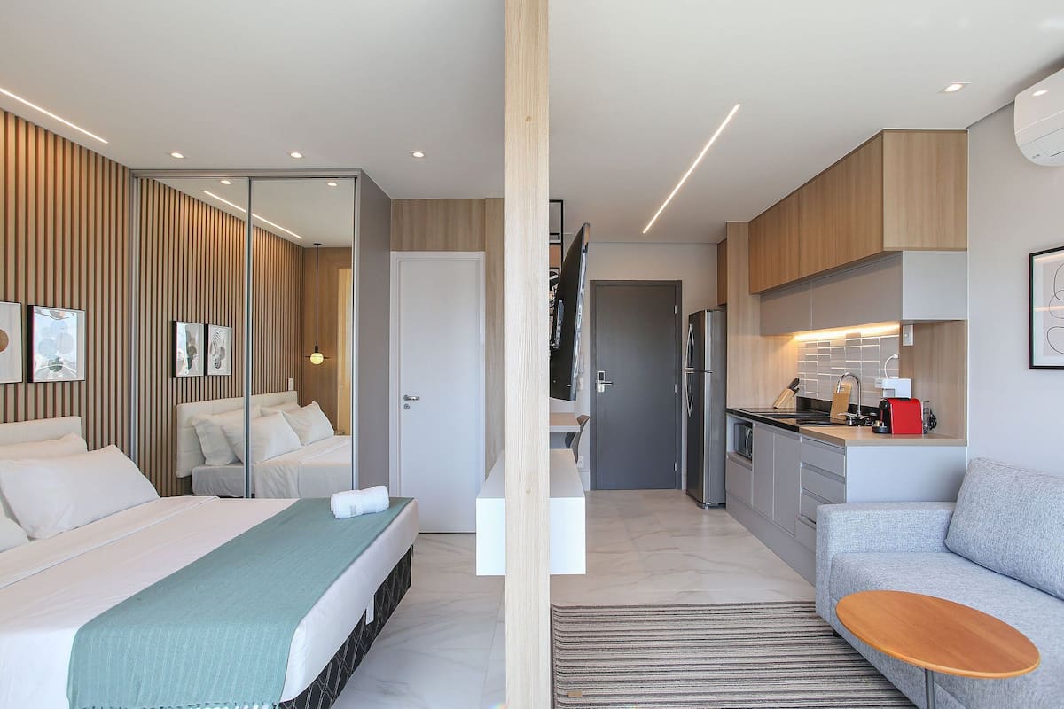 A modern hotel room with a bed, a sofa, a small table, a kitchen area, and a bathroom.