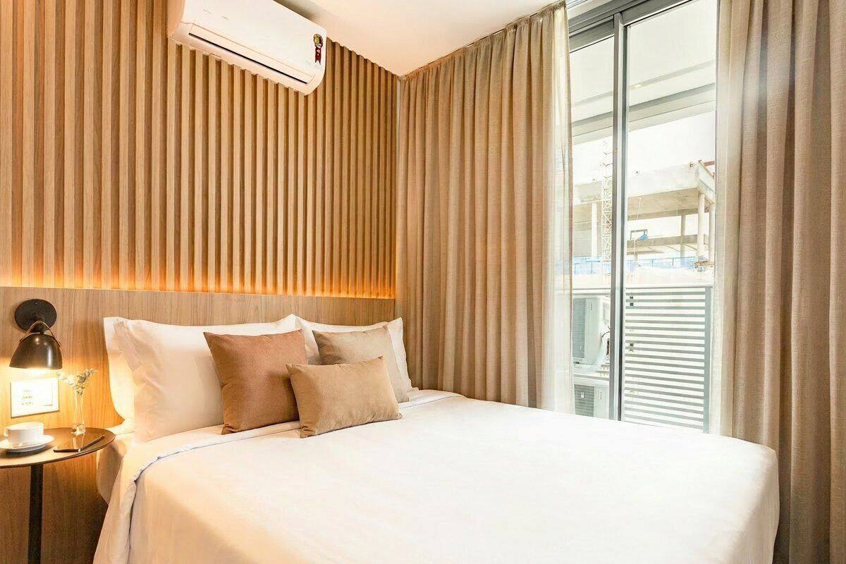 A hotel room with a bed, a nightstand with a lamp, a window with curtains, and a view of the outside.