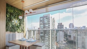 A balcony with a city view, a dining table with plates and glasses, and a green ottoman. - Helbor Wide (São Paulo)