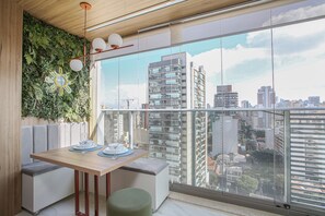 A balcony with a city view, a dining table with plates and glasses, and a green ottoman. - Helbor Wide (São Paulo)