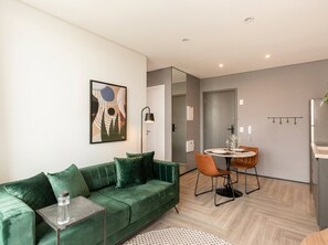 A modern living room with a green sofa, a small table with a water bottle, a dining area with a round table and chairs, and a kitchen area with a refrigerator.