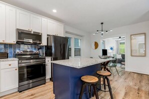 A modern kitchen with white cabinets, stainless steel appliances, a blue island with stools, and a dining area with a TV and seating.